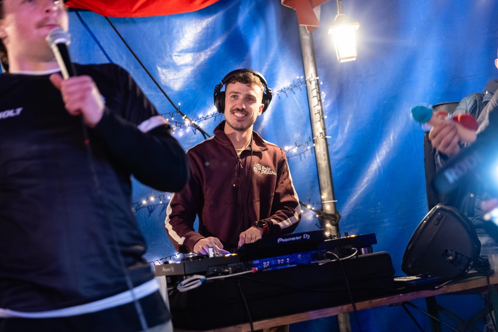 Mathis aux platines sous un chapiteau, casque sur les oreilles, pendant qu’un animateur parle au micro
